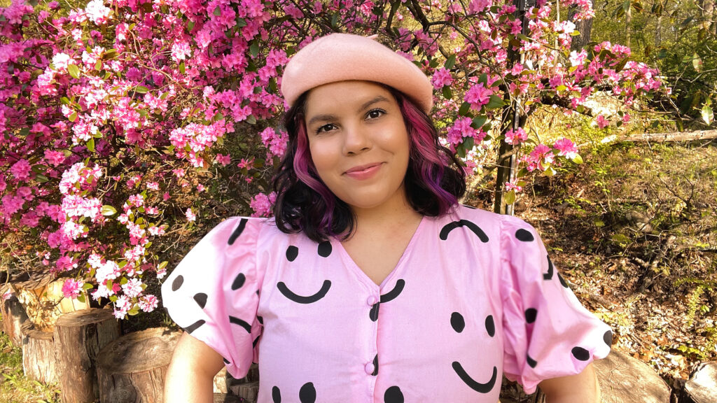 Author Claribel Ortega, photo credit Claribel A. Ortega. Photo shows a woman in a pink beret with a pink blouse with smiley faces on it sitting in front of a tree full of blossoming pink flowers.