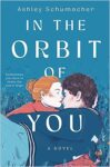 In the Orbit of You by Ashley Schumacher cover shows a girl with reddish hair in a bun wearing a turtleneck sweater kissing a boy's nose. The boy is lying in the grass wearing a jacket and jeans.